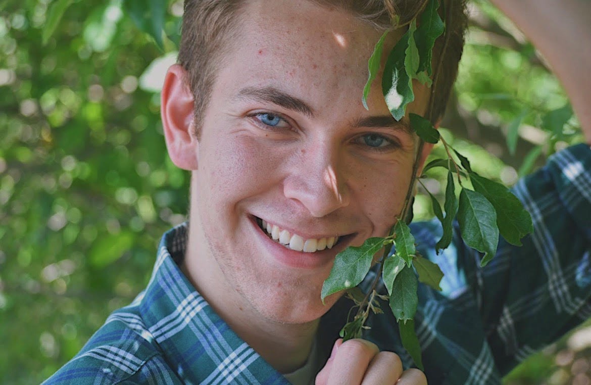 A young man poses for a photo wearing a blue button-up shirt next to a tree.