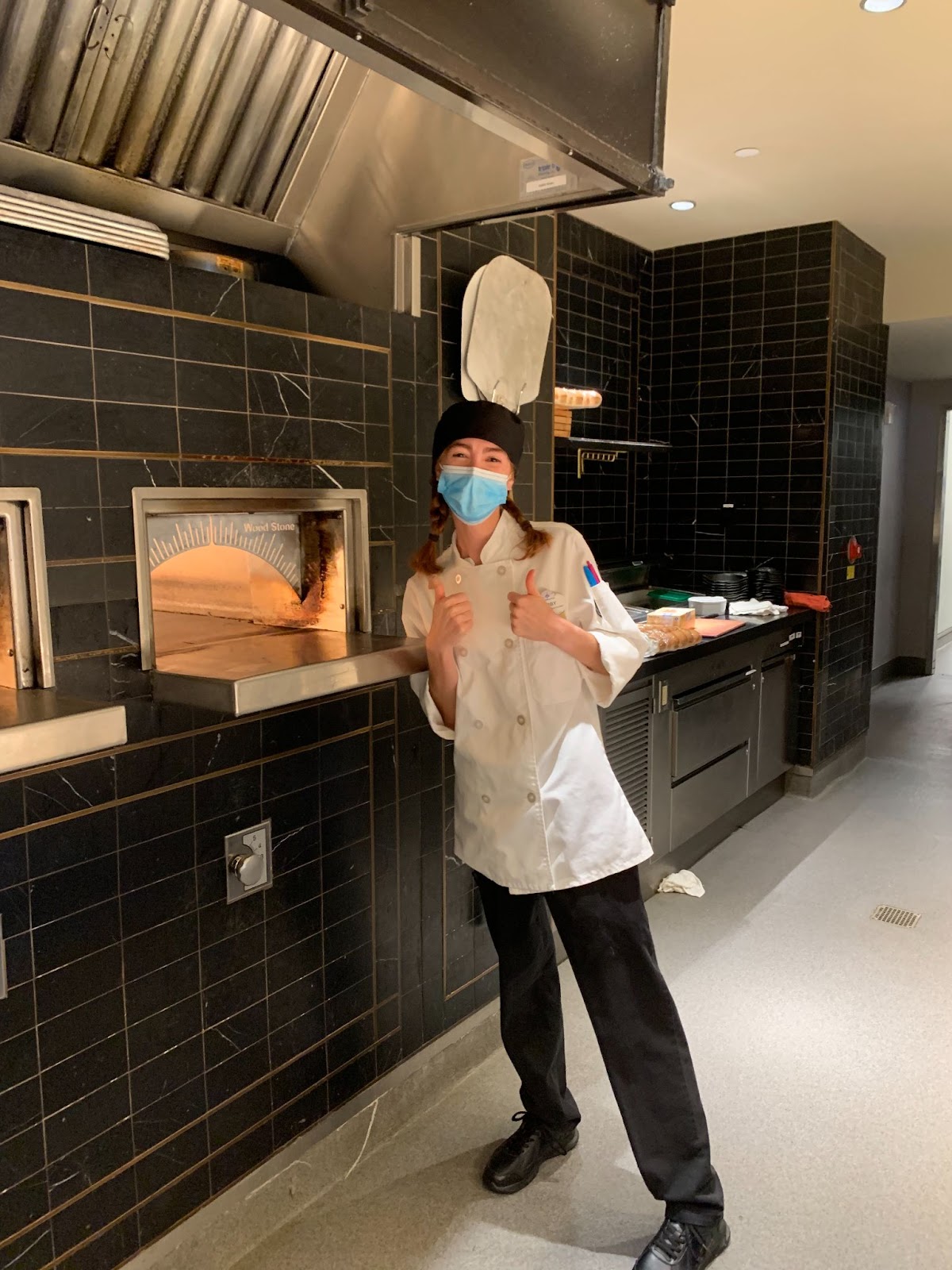 A woman in a chef apron smiling with thumbs up in front of an oven.